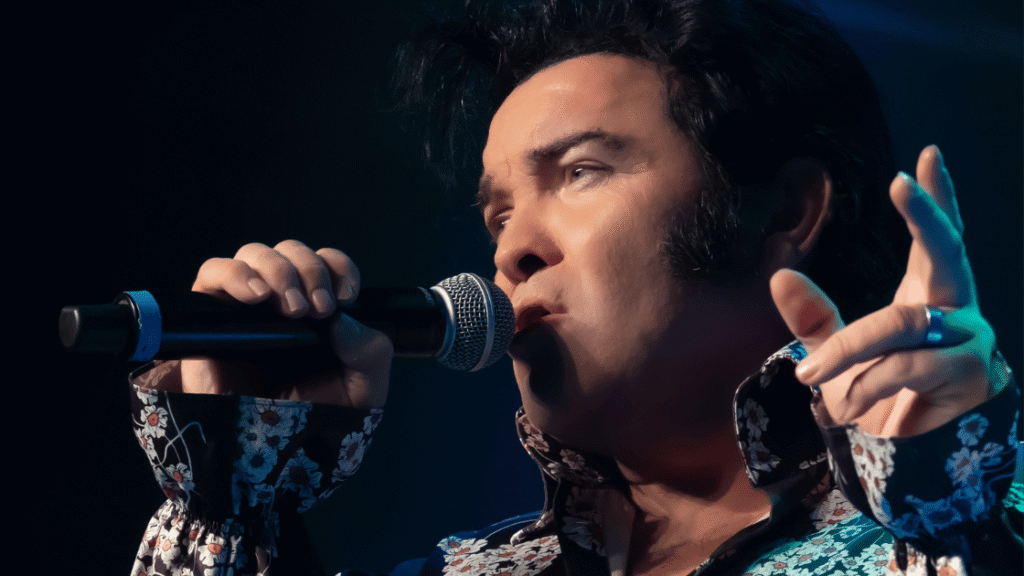 Close-up of a male singer performing on stage, holding a microphone and gesturing with his hand while singing.