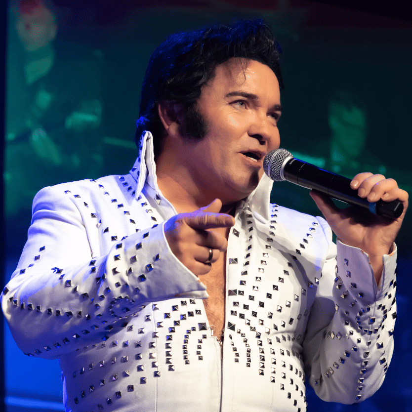 Elvis impersonator in a white studded jumpsuit sings into a handheld microphone on stage.