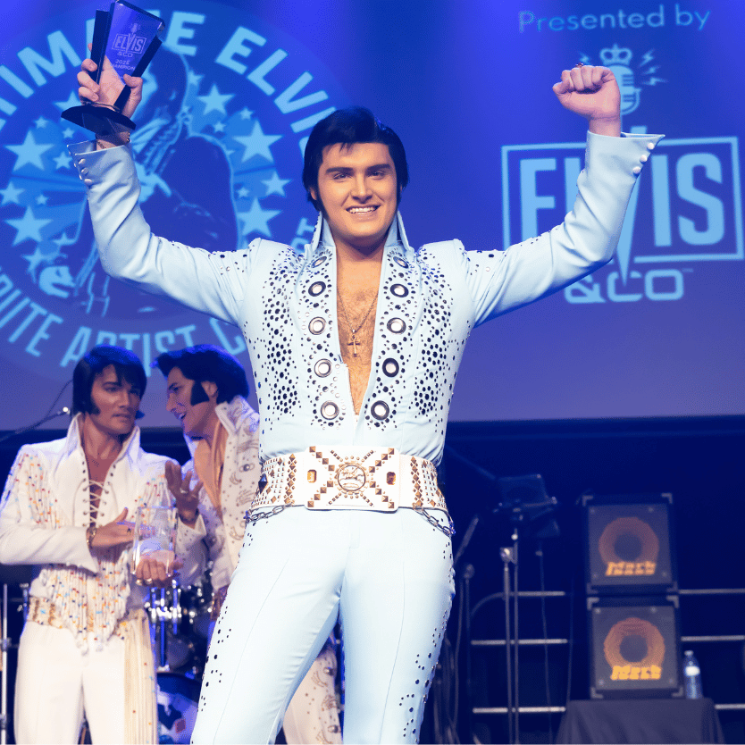 Elvis tribute artist on stage raising a trophy in his right hand, wearing a light-blue rhinestone-studded jumpsuit and belt.