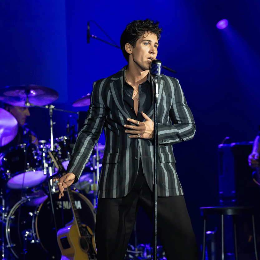 Male singer performing on stage with striped blazer, holding microphone stand and touching chest as he sings under blue stage lights.