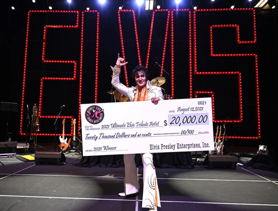 Elvis tribute artist on stage holding a large check for $20,000, with red ELVIS signs in the background.