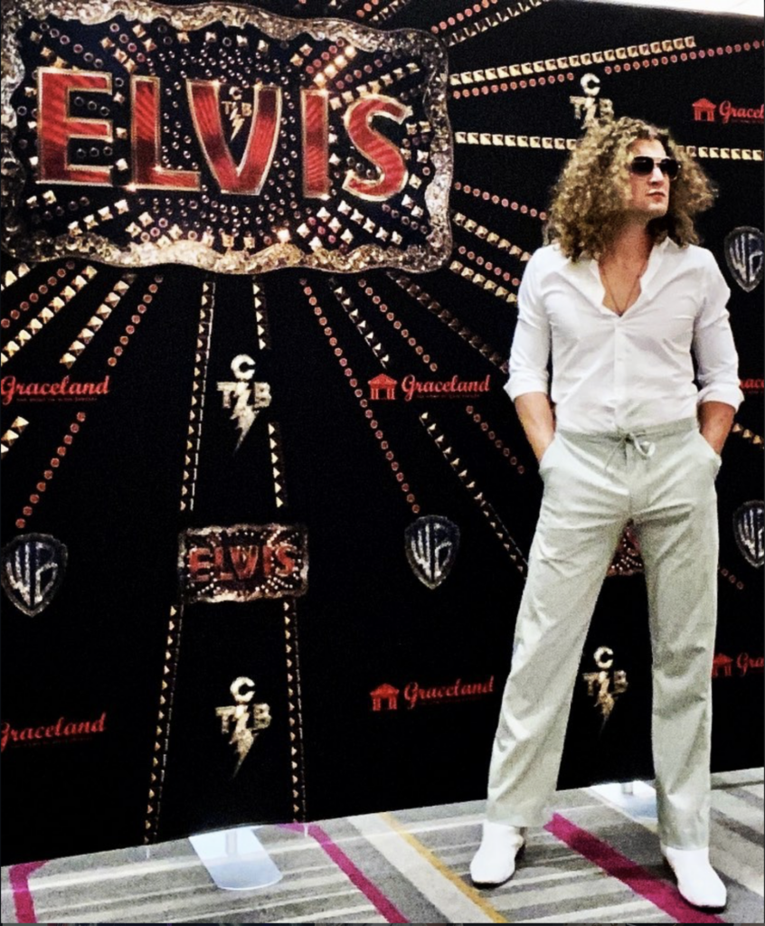 Man with long hair in a white shirt and light pants posing in front of a black, jeweled backdrop that says Elvis and Graceland logos.
