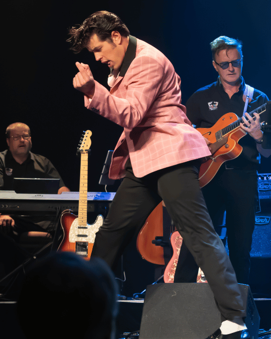 Male singer in a pink blazer performing on stage with a guitarist in the background; keyboard player visible on left and guitars around.