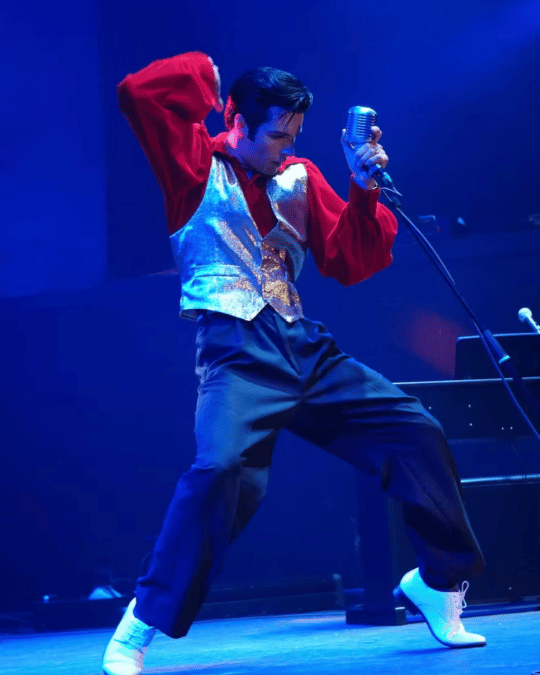 Male singer performing on stage with a vintage silver vest, red shirt, and white shoes under blue lighting, holding a retro microphone.