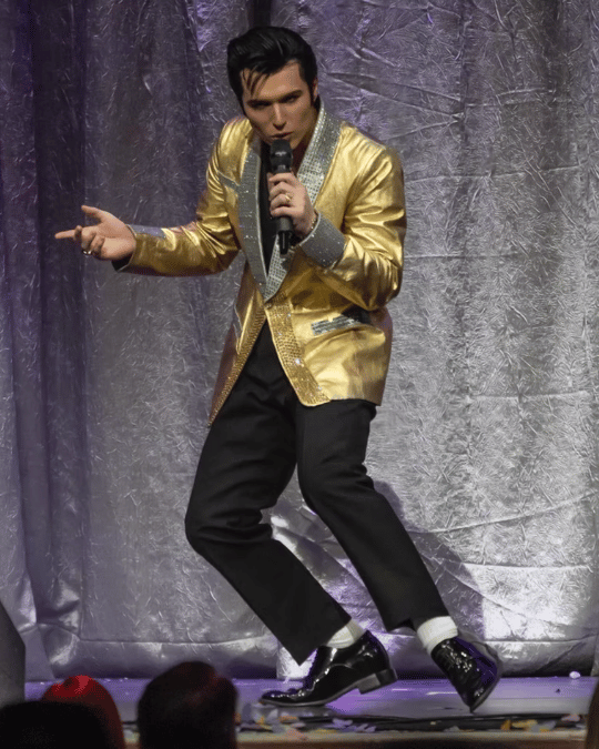 Male singer on stage wearing a shiny gold jacket and black pants, singing into a microphone while gesturing to the audience.