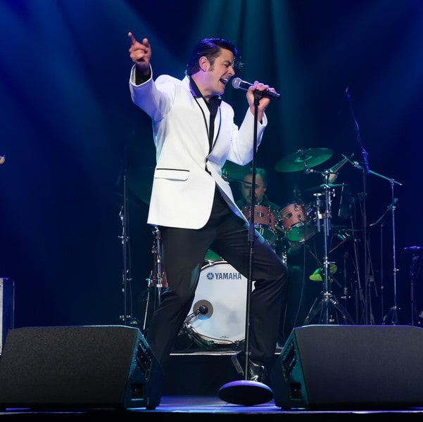 Male singer in a white blazer performs on stage with a microphone, drum set visible in the background under blue stage lights