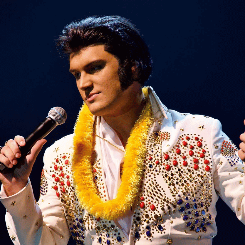 Elvis impersonator performing on stage, holding a microphone, wearing a white rhinestone jumpsuit and a yellow lei.