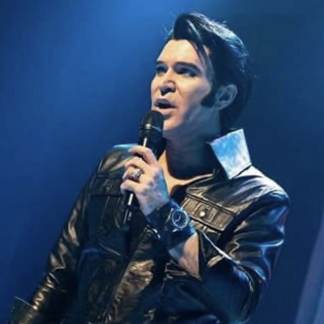Male singer in a black leather jacket sings into a handheld microphone on stage under blue lighting.