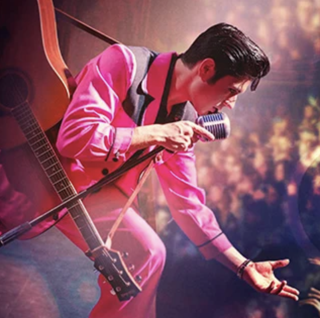 Elvis-style performer in a pink suit singing into a microphone on stage with a guitar strapped nearby.