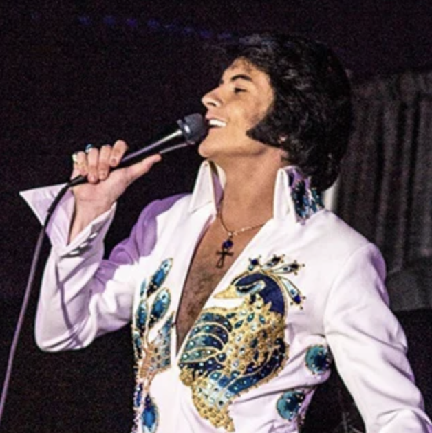 Male Elvis Presley tribute artist singing into a microphone on stage, wearing a white jumpsuit with blue/gold embroidery and an open chest area.