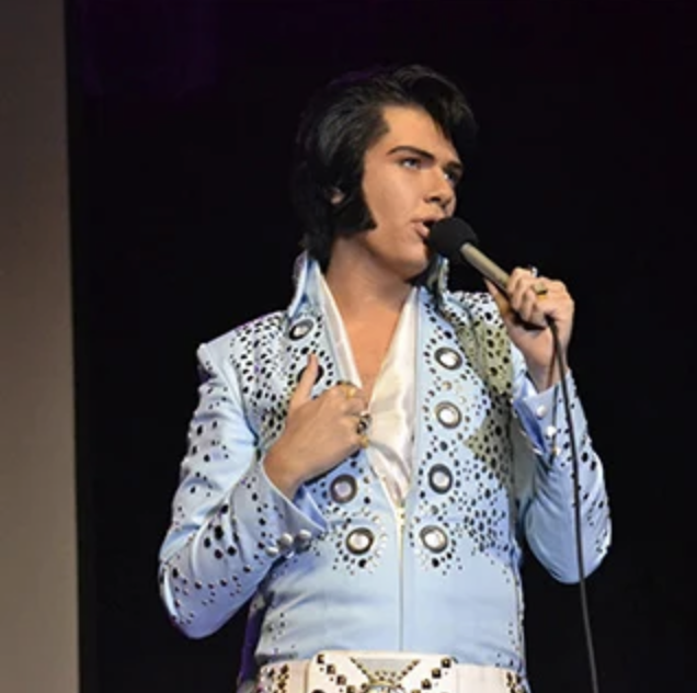 Elvis impersonator on stage, wearing a light blue rhinestone jumpsuit and singing into a microphone.