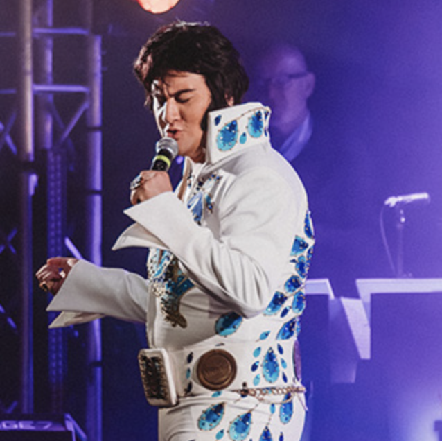 Elvis tribute artist singing into a microphone on stage in a white jeweled jumpsuit with blue accents.