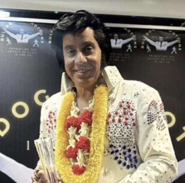 Elvis Presley look-alike in a rhinestone white jumpsuit, wearing yellow and red flower garlands, holding a glass trophy at an awards ceremony.