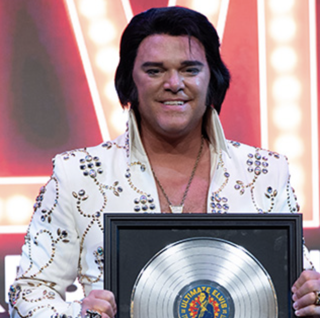Man wearing a white studded jacket holds a framed platinum record, posing for a photo on a stage backdrop with bright lights