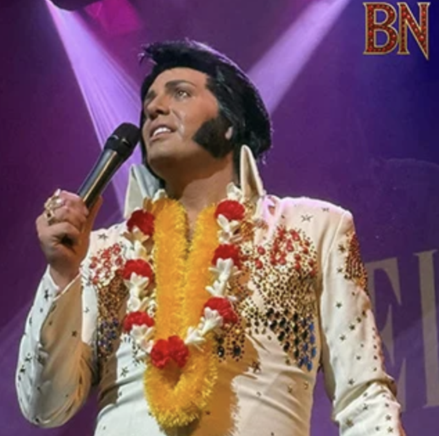 Elvis impersonator on stage in a jeweled white jumpsuit with yellow and red leis, holding a microphone. BN logo appears in the top-right corner.