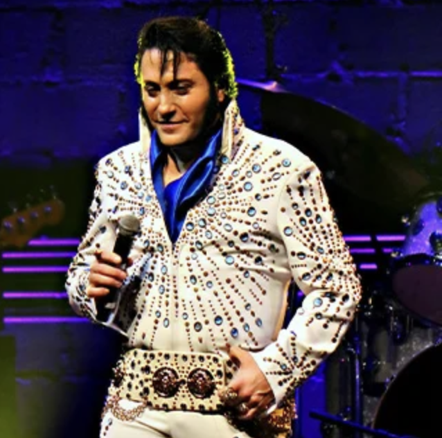 Elvis impersonator in a white gem-studded jumpsuit holding a microphone on stage.
