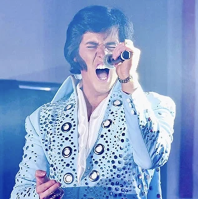 Male Elvis-style performer singing into a handheld microphone on stage, wearing a light blue studded jumpsuit.