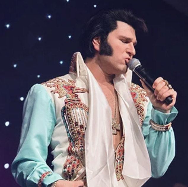 Elvis Presley look-alike singer performing on stage in a turquoise and white embellished jumpsuit, holding a microphone.
