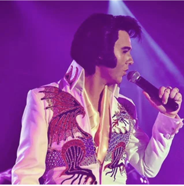 Elvis impersonator in a white jumpsuit with dragon embroidery singing into a microphone on a purple-lit stage