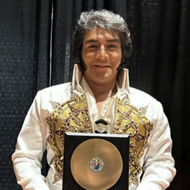 Elvis impersonator in a white jumpsuit with gold embroidery holding a gold record plaque behind black backdrop.