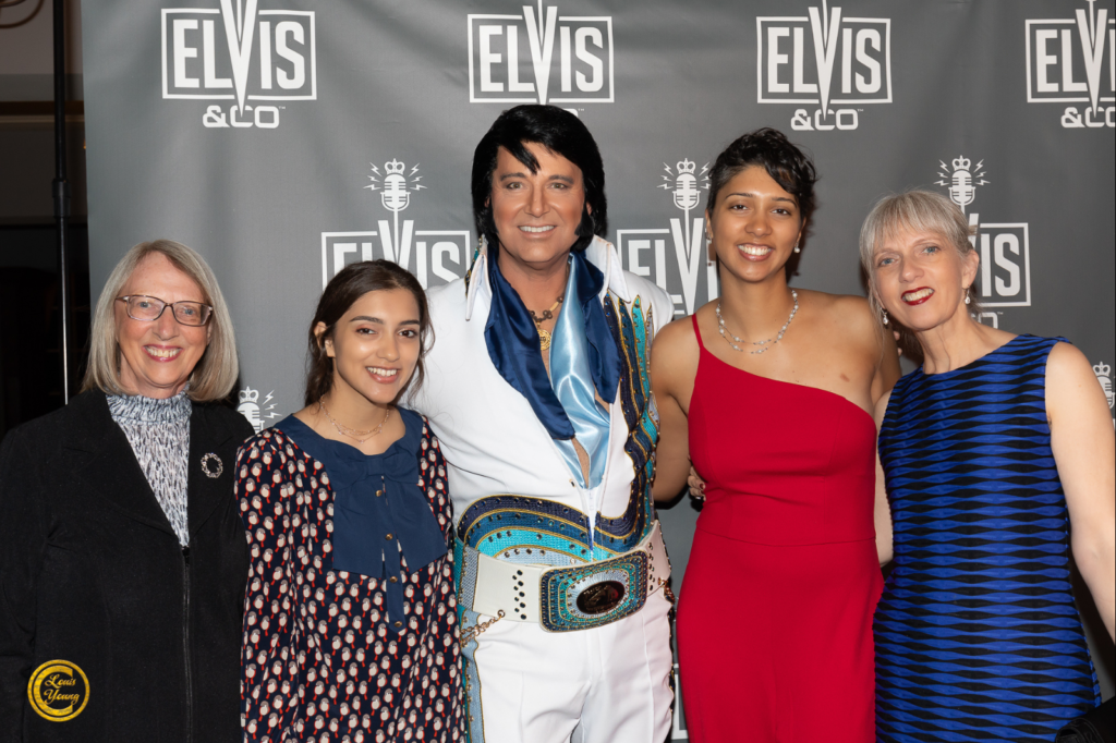 Five people pose together in front of an Elvis & Co backdrop at an event; center figure is an Elvis impersonator in a white jumpsuit with blue scarf and jeweled belt.