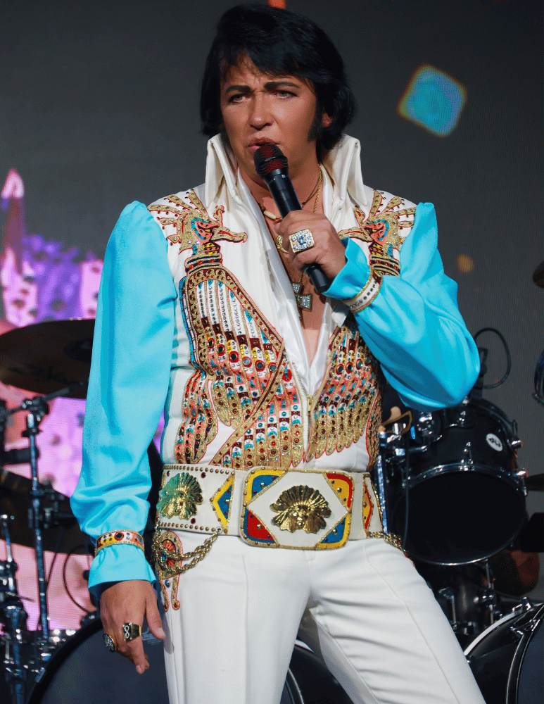 Elvis impersonator on stage singing into a microphone, wearing a white jumpsuit with blue sleeves and jeweled embellishments.