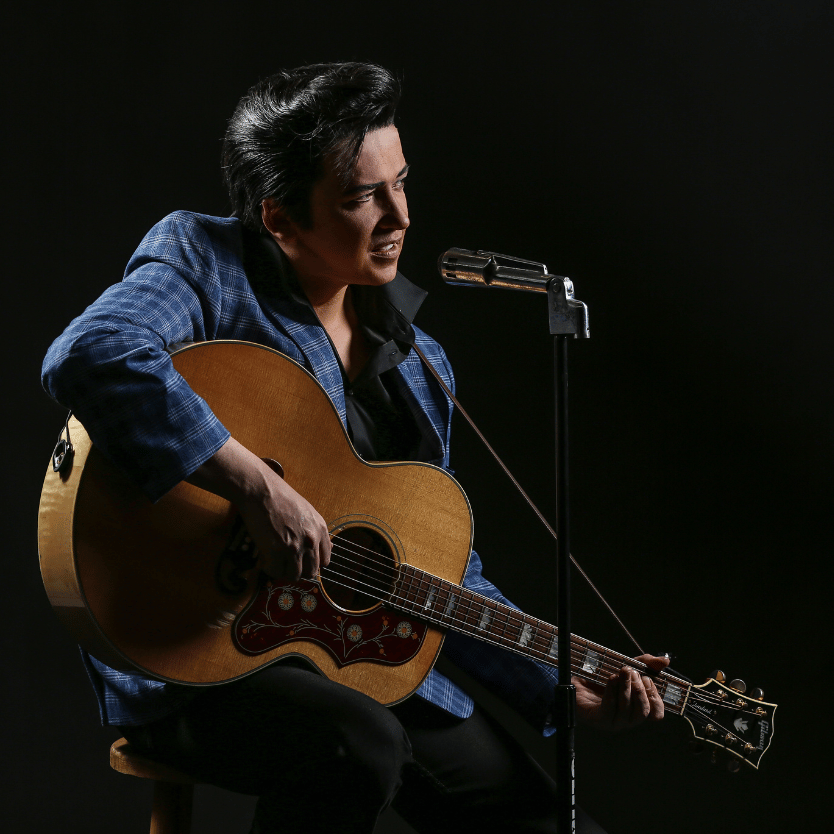 Musician seated on a stool playing an acoustic guitar with a vintage design, singing into a microphone against a dark backdrop.