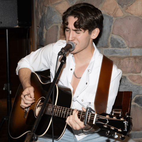 Man in a white shirt sings into a microphone while playing an acoustic guitar on a stone-wall stage backdrop.